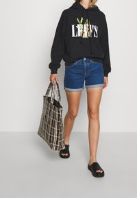 Un sweat à capuche noir avec le logo "Levi's", un short en denim bleu, des sandales noires et un sac fourre-tout à motifs vichy dans des tons bruns et crème.