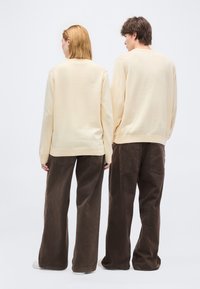 Deux personnes se tenant dos à la caméra, portant des pulls crème, des pantalons marron à jambes larges et des chaussures blanches sur un fond blanc uni.