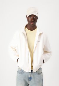 Hvid zip-up jakke med elastiske manchetter, en lys gul t-shirt under og lyseblå jeans. Beige kasket med logo. Enkel design og farver.