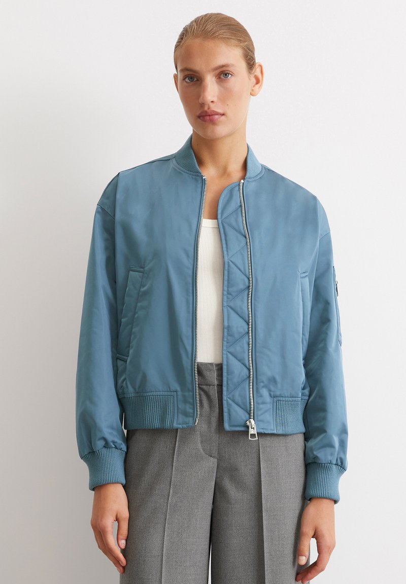 Jasnoniebieska kurtka bomberka wykonana z gładkiego materiału, z zamkiem błyskawicznym z przodu, prążkowanymi mankietami, bocznymi kieszeniami i lekko skróconym krojem.