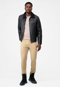 Black leather jacket with a shearling collar, beige shirt, tan pants, and black combat boots, model standing against a white background.