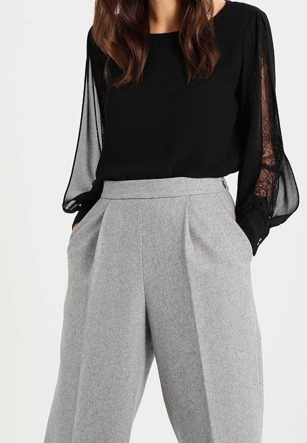 Femme portant un chemisier noir à manches longues avec des inserts en dentelle et un pantalon gris ample, taille haute, avec des poches.