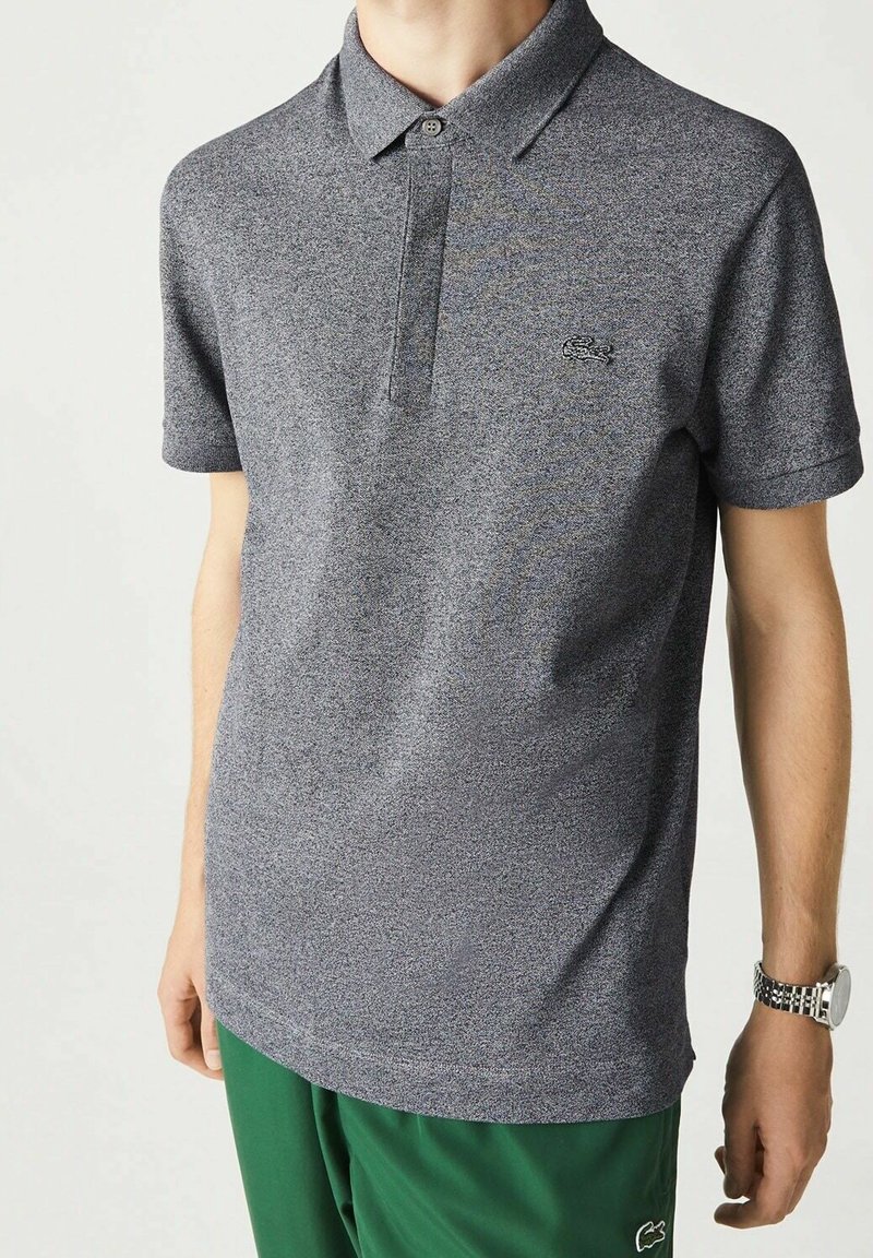 Homme portant un polo gris chiné à manches courtes avec un petit logo sur la poitrine et un pantalon vert, montre au poignet gauche.