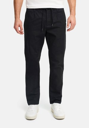 Mężczyzna w czarnych spodniach na sznurki i białych trampkach na jednolitym jasnym tle, stoi z ramionami rozluźnionymi.