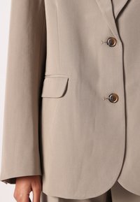 Beige blazer med et notched revers, enkelt knappelukking og en lomme. Stoffet er glatt med en matt finish.