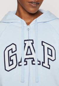 Primer plano de una persona que lleva una sudadera de cremallera azul claro con "GAP" bordado en grandes letras blancas contorneadas en un tono azul marino.