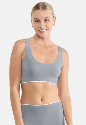 Uśmiechnięta kobieta w jasnoszarym sportowym staniku i dopasowanych majtkach z wysokim stanem na jednolitym białym tle.