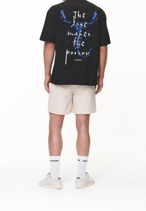 Zwart T-shirt met een blauw-wit grafisch ontwerp op de achterkant, gecombineerd met beige korte broek, witte sokken en lichte sneakers.
