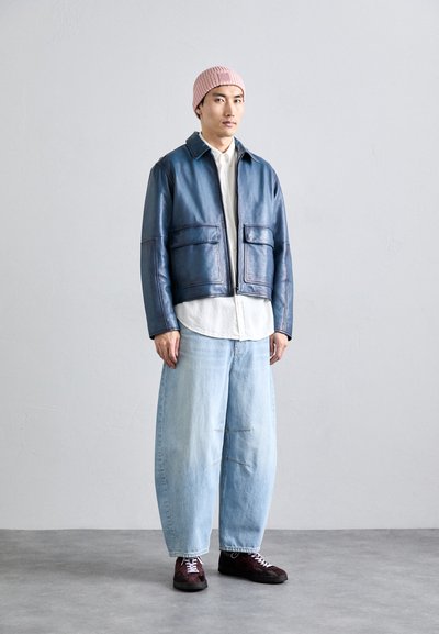 Man in een roze gebreide beanie, blauwe leren jas, wit shirt, wijde lichtblauwe jeans en donkere sneakers op een grijze vloer.