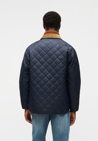 Μπλε καπιτονέ μπουφάν με μοτίβο ρόμβων, γιακά από κοτλέ σε καφέ απόχρωση, άνετη γραμμή και πλαϊνές τσέπες. Προβολή από πίσω που δείχνει ομαλή υφή.