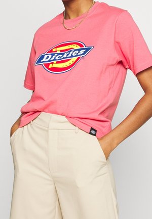 Henkilö, joka pukeutuu vaaleanpunaiseen Dickies-logoiseen T-paitaan, joka on sujautettu beigeihin housuihin, kädet taskuissa ja kullanvärinen ketjukoru kaulassa.