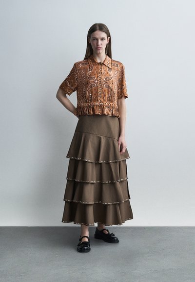 Νέα γυναίκα που στέκεται με το χέρι στη μέση, φορώντας πορτοκαλί μπλούζα με σχέδια και κοντά μανίκια και καφέ φούστα με βαθμίδες, μαζί με μαύρα παπούτσια.