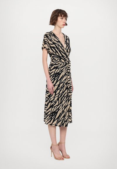 Vestido hasta la rodilla en beige con un patrón abstracto negro, con escote en V, mangas cortas y detalle de cintura anudada. Usado con tacones con correas.