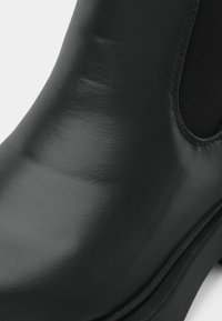 Czarne botki sięgające kostki o błyszczącej powierzchni, z elastycznymi panelami bocznymi i gładkim, zaokrąglonym noskiem. Posiadają grubą czarną podeszwę o subtelnej teksturze.