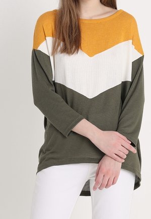 Vrouw draagt een langarmig topje met kleurvlakken in mosterdgeel, wit en olijfgroen, gecombineerd met een witte broek, poseert met over elkaar geslagen handen.
