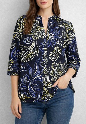 Donna che indossa una camicetta blu navy con bottoni e motivi floreali gialli e azzurri, abbinata a jeans blu, una mano in tasca.