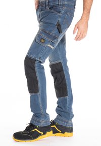 Pantalons de travail en denim bleu avec des pare-knees renforcés en noir, plusieurs poches et un design slim fit. Coutures contrastées et tissu durable.