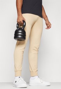 Beige tapered sweatpants made of soft fabric, featuring ribbed cuffs and side seams. A person holds a black kettlebell in one hand.