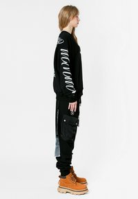 Sudadera negra de gran tamaño con gráficos blancos, combinada con pantalones joggers negros que presentan un gran bolsillo. Botas marrones con suelas gruesas.