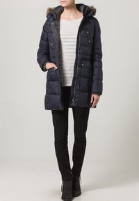 Giubbotto imbottito blu navy con cappuccio bordato di pelliccia, chiusura con zip e diverse tasche con automatico, indossato sopra una maglietta a righe e pantaloni neri.