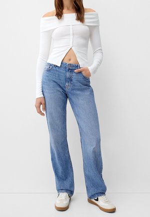 Bílý crop top s odhalenými rameny a dlouhými rukávy, doplněný o knoflíkovou detail, v kombinaci se světle modrými kalhotami s širokými nohavicemi a bílými teniskami s hnědými podrážkami.