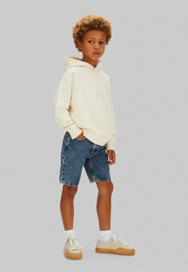 Niño con cabello rizado que lleva una sudadera con capucha color crema, pantalones cortos de mezclilla, calcetas blancas y zapatillas beige, de pie contra un fondo gris liso.