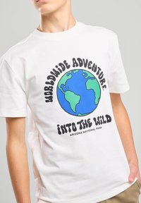 Camiseta blanca de algodón con un gráfico multicolor de la Tierra y el texto "Aventura Mundial en la Naturaleza" debajo.