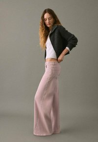 Jeans de pierna ancha rosa claro con botones laterales, combinados con una blusa blanca ajustada y un cárdigan oscuro de textura. Atuendo simple y casual.