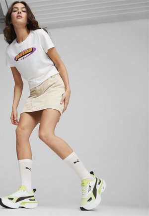 T-shirt bianco con logo viola e arancione, gonna in corda color beige, calze bianche con logo nero e scarpe chunky gialle e bianche.