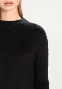 Primo piano di una persona che indossa un maglione in maglia nera con scollatura a girocollo su uno sfondo chiaro e uniforme.