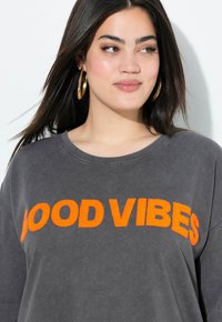 Sudadera gris de gran tamaño con el texto bordado "BUENAS VIBRAS" en naranja. Presenta un cuello redondo y mangas largas. Textura de tela suave.