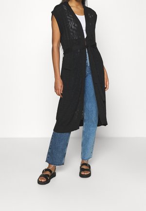 Femme portant un cardigan en maille noir sans manches avec ceinture, un haut blanc, un jean bleu et des sandales noires, debout sur fond blanc.
