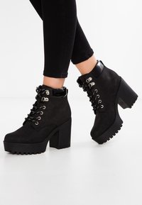 Black ankle boots made of suede with a matte finish. Features a thick block heel, silver lace-up hardware, and glossy black accents.
