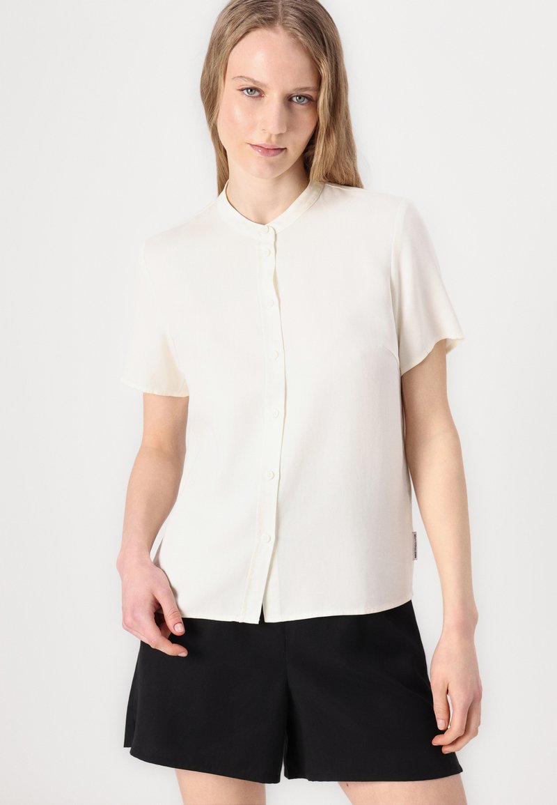 Helles, cremefarbenes Kurzarmblusenshirt mit Kragen, ausgestattet mit einer durchgängigen Knopfleiste und einem glatten, leichten Stoff. Kombiniert mit schwarzen Shorts.