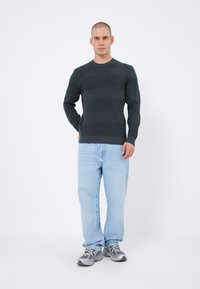 Un pull texturé vert foncé avec des motifs de rayures horizontales, associé à un jean droit bleu clair et des chaussures de sport grises.