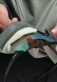 Schwarze Sonnenbrille mit einem Farbverlauf-Glas wird in einen hellgrauen, strukturierten Stoffbeutel gelegt. Der Beutel hat ein weiches Futter.
