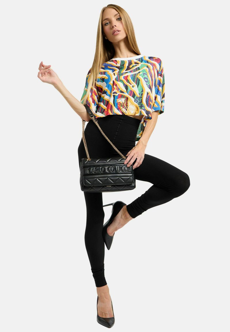 Kleurrijk, patroon crop top, zwarte legging en een zwart gewatteerde handtas met kettingriem. Model draagt zwarte hoge hakken en poseert op één been.