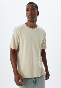 Beige t-shirt met korte mouwen in een losse pasvorm, gemaakt van zachte stof, met een ronde halslijn en subtiele tekstdetail "TIMELESS" op de borst.
