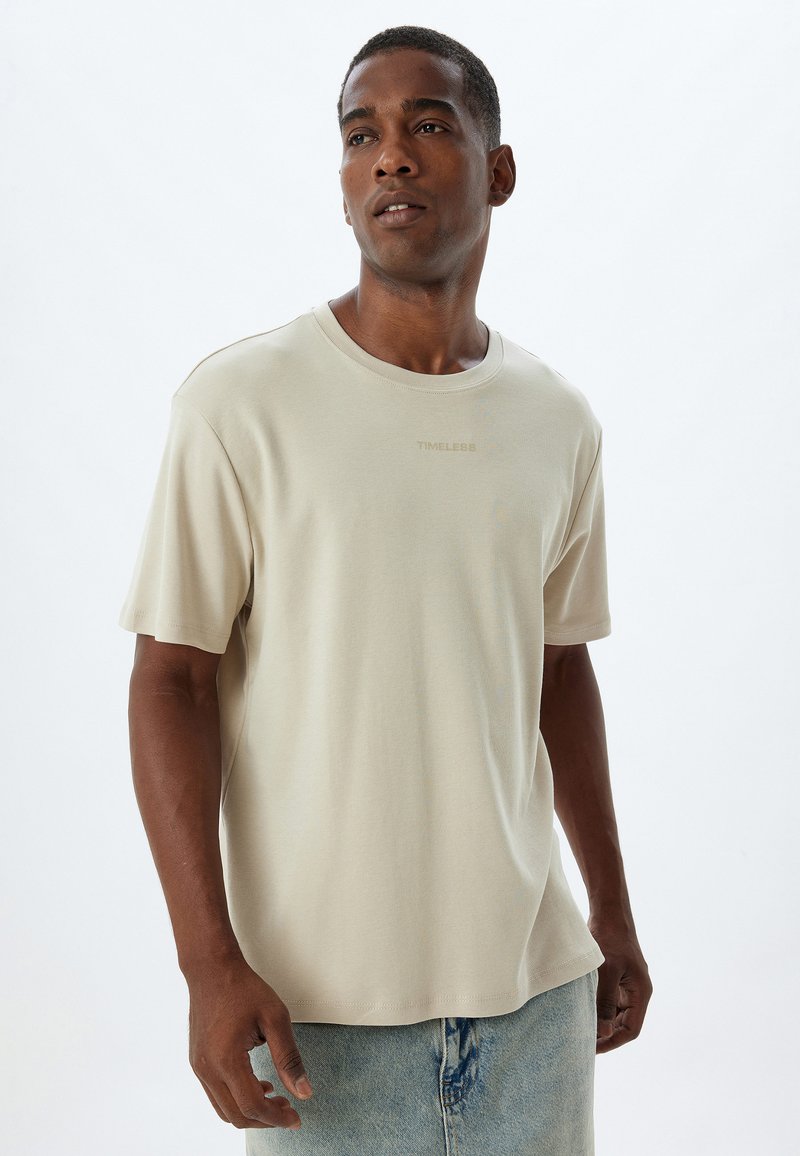 Beige t-shirt met korte mouwen in een losse pasvorm, gemaakt van zachte stof, met een ronde halslijn en subtiele tekstdetail "TIMELESS" op de borst.