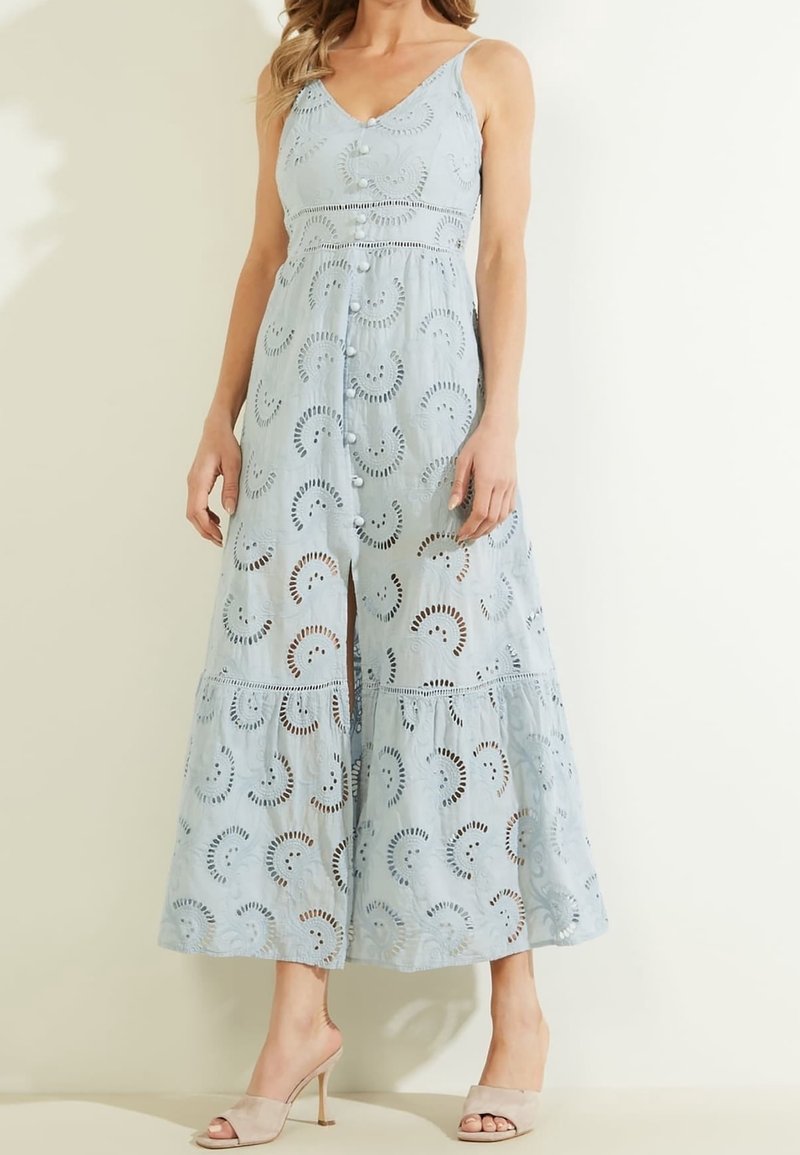 Femme portant une robe midi sans manches bleu clair avec des boutons sur le devant et des motifs circulaires en broderie anglaise, associée à des talons ouverts beige.