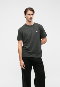 Ung mann iført en mørkegrå T-skjorte med en liten logo og svarte jeans, stående med en hånd bak ryggen mot en ensfarget bakgrunn.