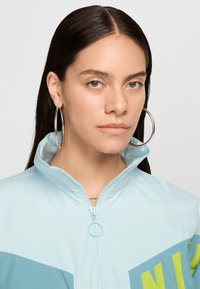 Jachetă cu fermoar de culoare albastră deschisă, cu guler înalt, având accente contrastante de albastru închis și verde lime și o textură netedă.