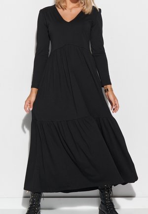 Kobieta w długiej czarnej sukience z falbanami, z długimi rękawami, dekoltem w serek, złotą bransoletką i czarnymi sznurowanymi botkami.