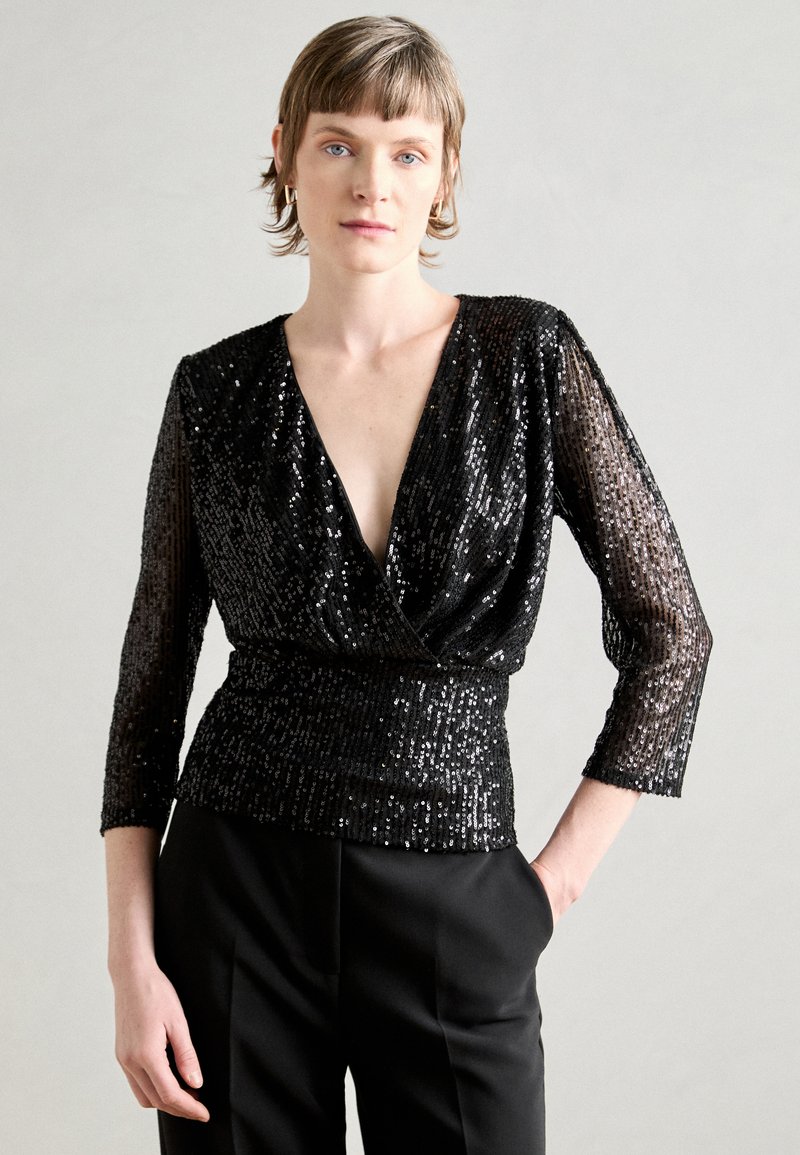 Schwarzes, mit Pailletten besetztes Oberteil mit tiefem V-Ausschnitt und drei Viertelärmeln. Texturiert, figurbetont in der Taille, kombiniert mit eleganten schwarzen Hosen.