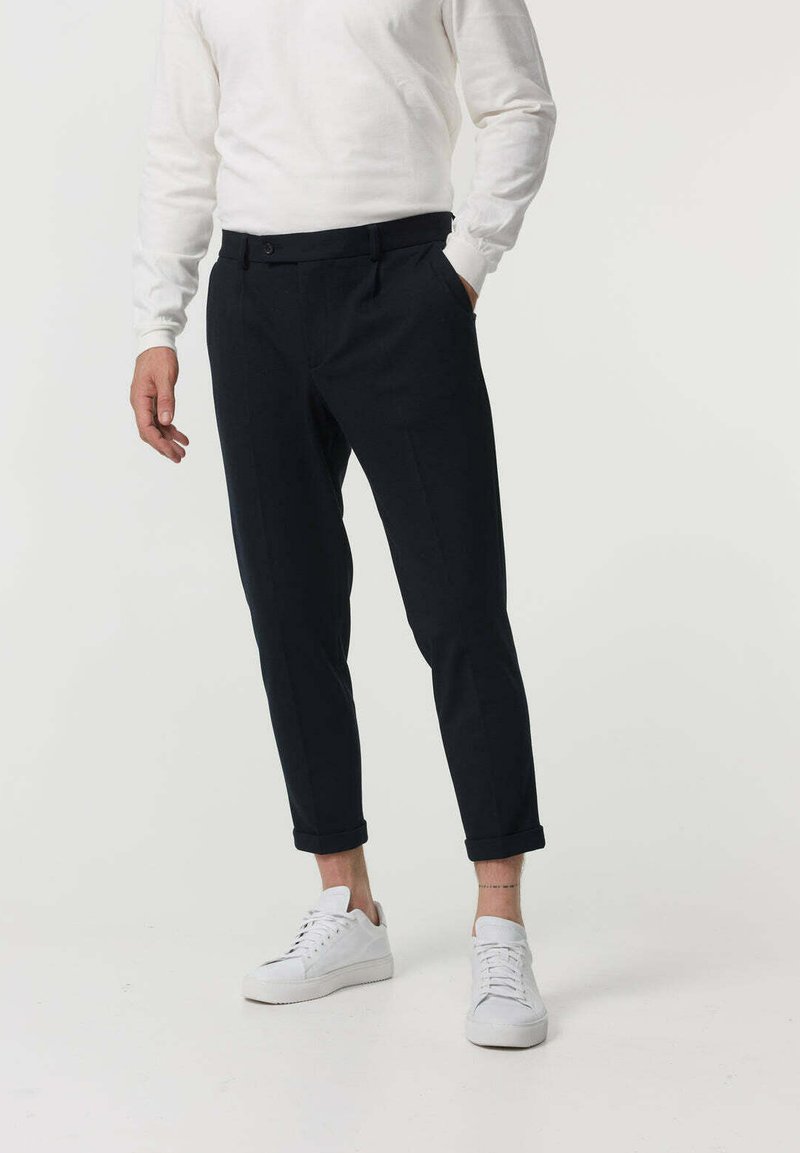 Pantaloni tailored de un albastru marin, cu un design cu picior îngustat, prezentând un bucluc pentru curea, închidere cu un singur nasture și tivuri răsucite, asortați cu adidași albi.