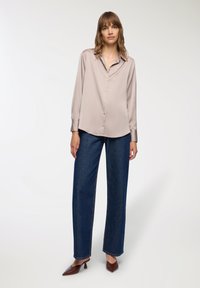 Camicia a maniche lunghe in satin beige chiaro con colletto a punta, chiusura con bottoni frontalmente e orlo leggermente curvo, abbinata a jeans scuri a gamba larga.