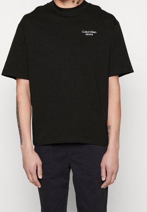 Osoba nosząca czarną koszulkę Calvin Klein Jeans z krótkimi rękawami i widocznymi tatuażami na przedramionach, w połączeniu z ciemnymi spodniami.