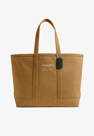 Sac fourre-tout marron Coach avec deux poignées, texte de logo blanc sur le devant et étiquette en cuir noir suspendue à la poignée droite.