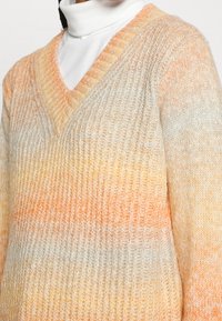 Pull en tricot avec un col en V, présentant un motif dégradé de teintes crème, orange et jaune clair. Des points texturés ajoutent de la profondeur.