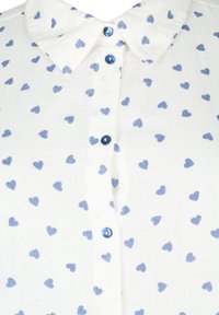 White shirt with a collar, featuring a pattern of small blue hearts. It has blue buttons along the front. Light, soft fabric texture.